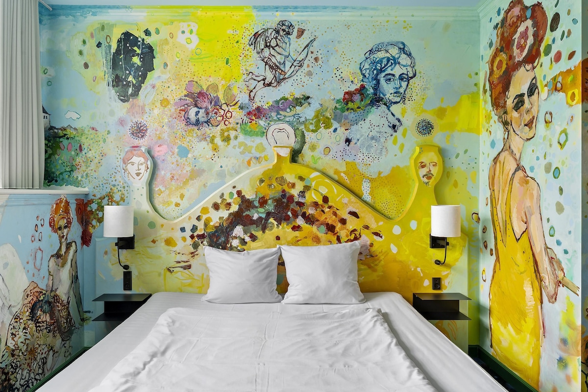 A hotel room with a bed, two bedside lamps, and a colorful abstract mural on the wall.