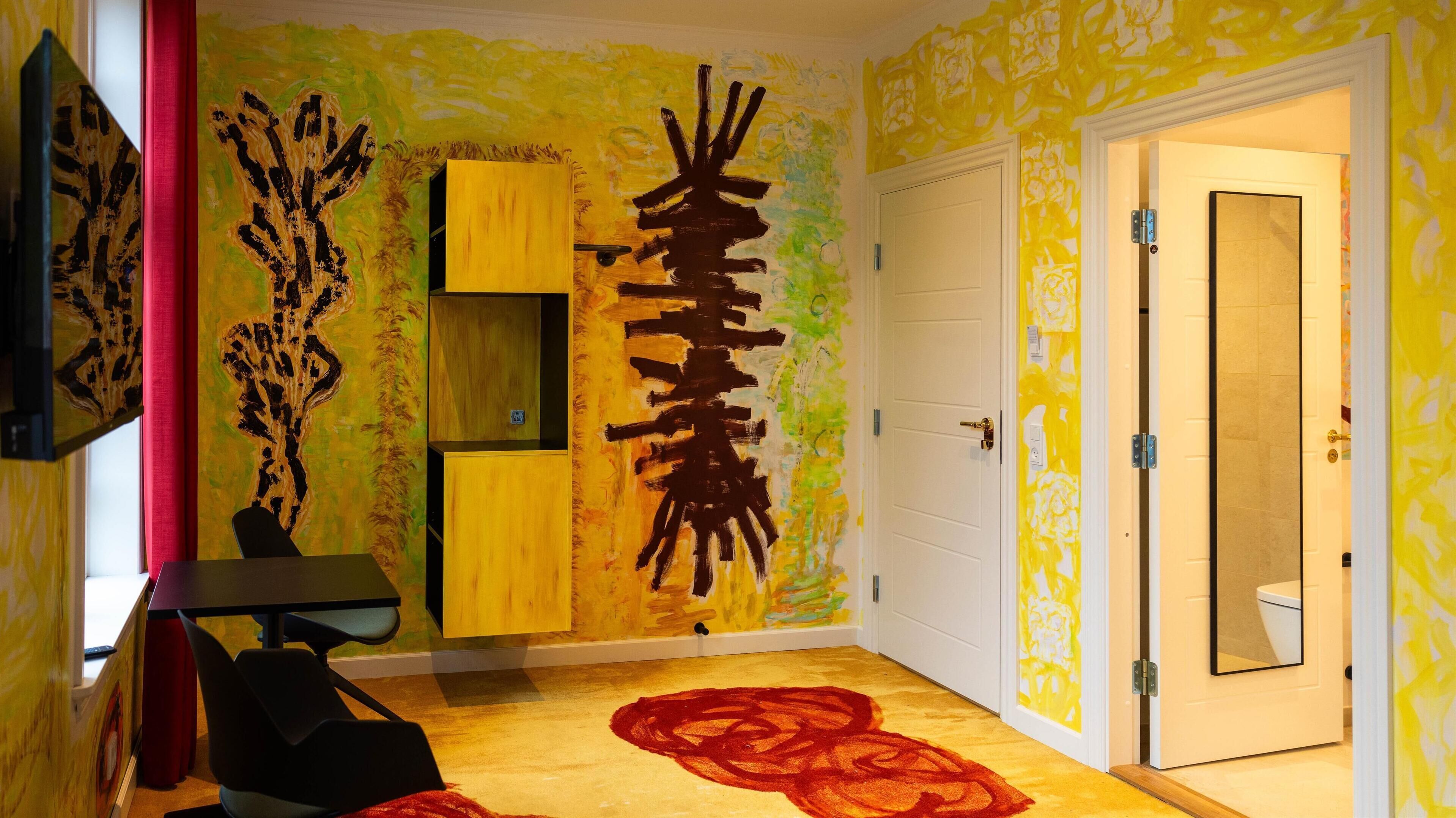 A room with a yellow and green abstract wall design, a wooden cabinet, a black chair, and a red rug.