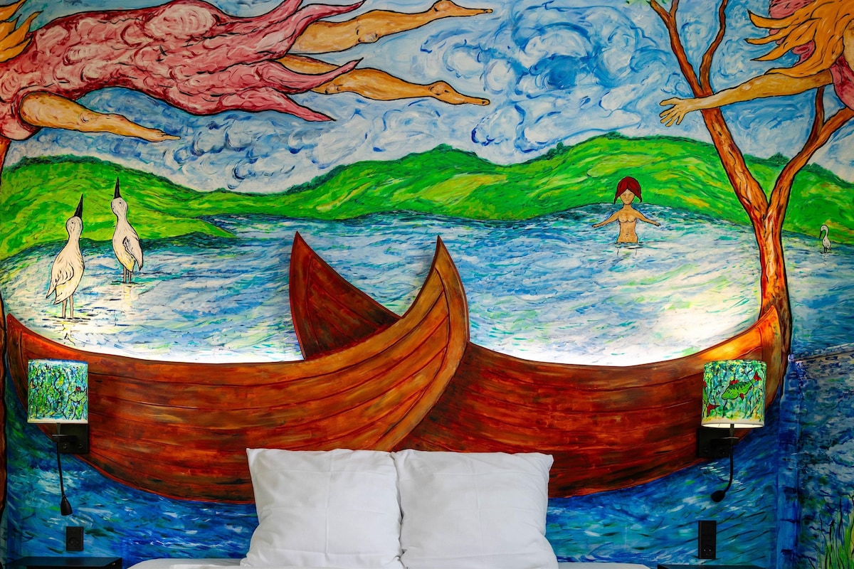 A bedroom with a mural of a river scene, a bed, and bedside tables.