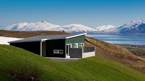 Een modern gebouw met een schuin dak, gelegen op een grasheuvel met op de achtergrond besneeuwde bergen en een wateroppervlak.