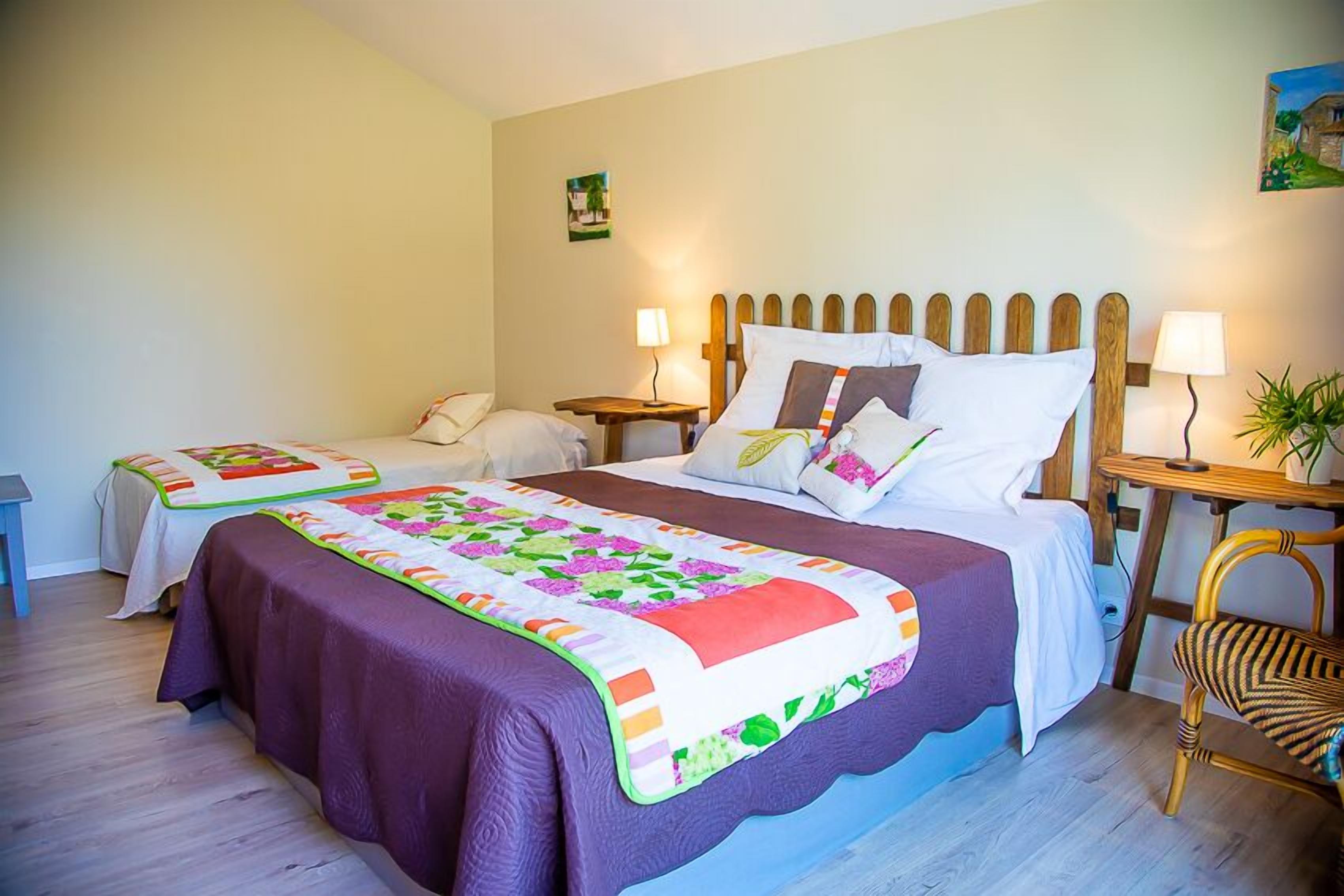 A hotel room with two beds, each with a wooden headboard, and a large bed with a colorful quilt. There are bedside tables with lamps and a small table with a plant.