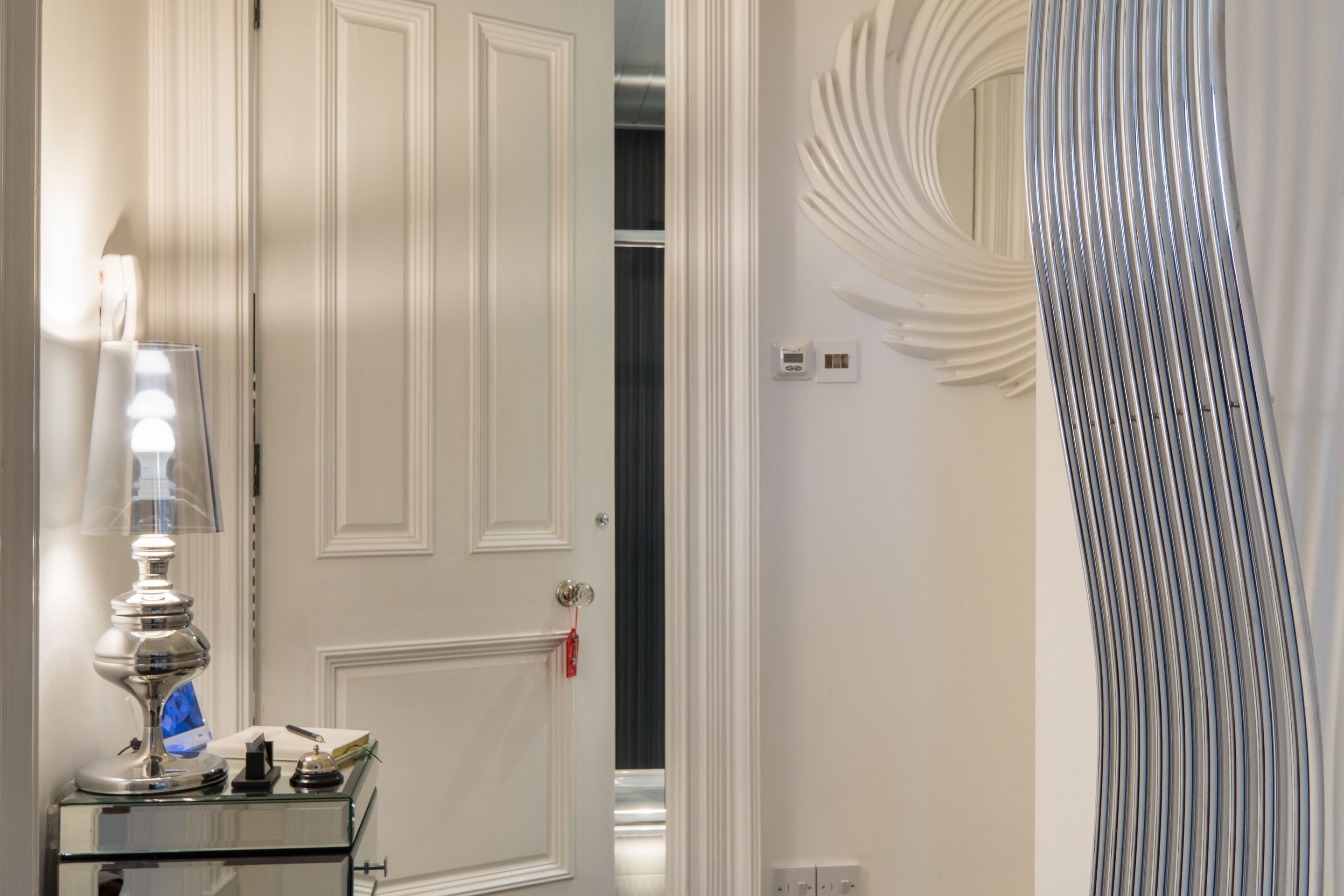 A hotel room with a closed door, a bedside table with a lamp, and a modern wall design.