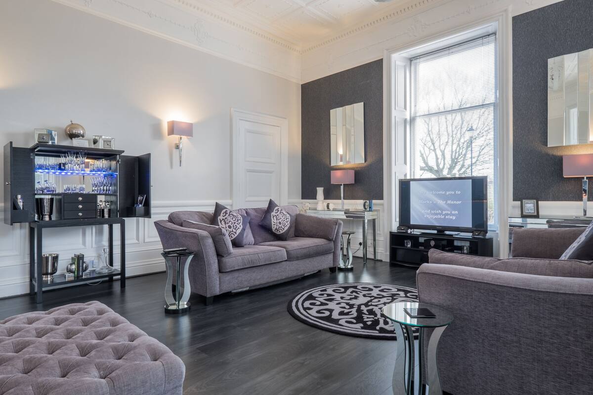 A modern living room with a grey sofa, a round coffee table, and a television. There is a decorative mirror on the wall and a small side table.