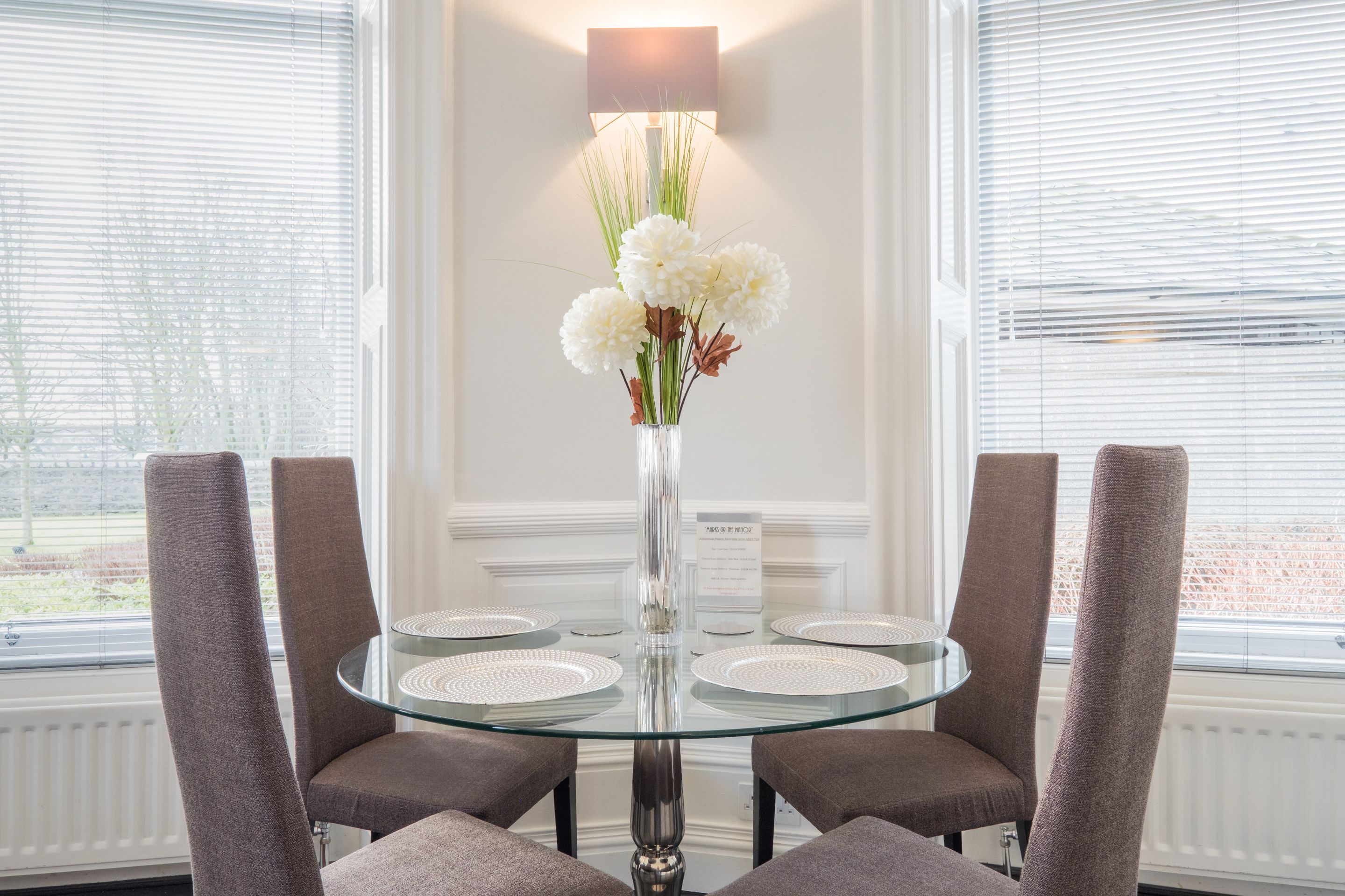 Una mesa redonda de vidrio con cuatro sillas, un florero con flores blancas y una lámpara en la pared.