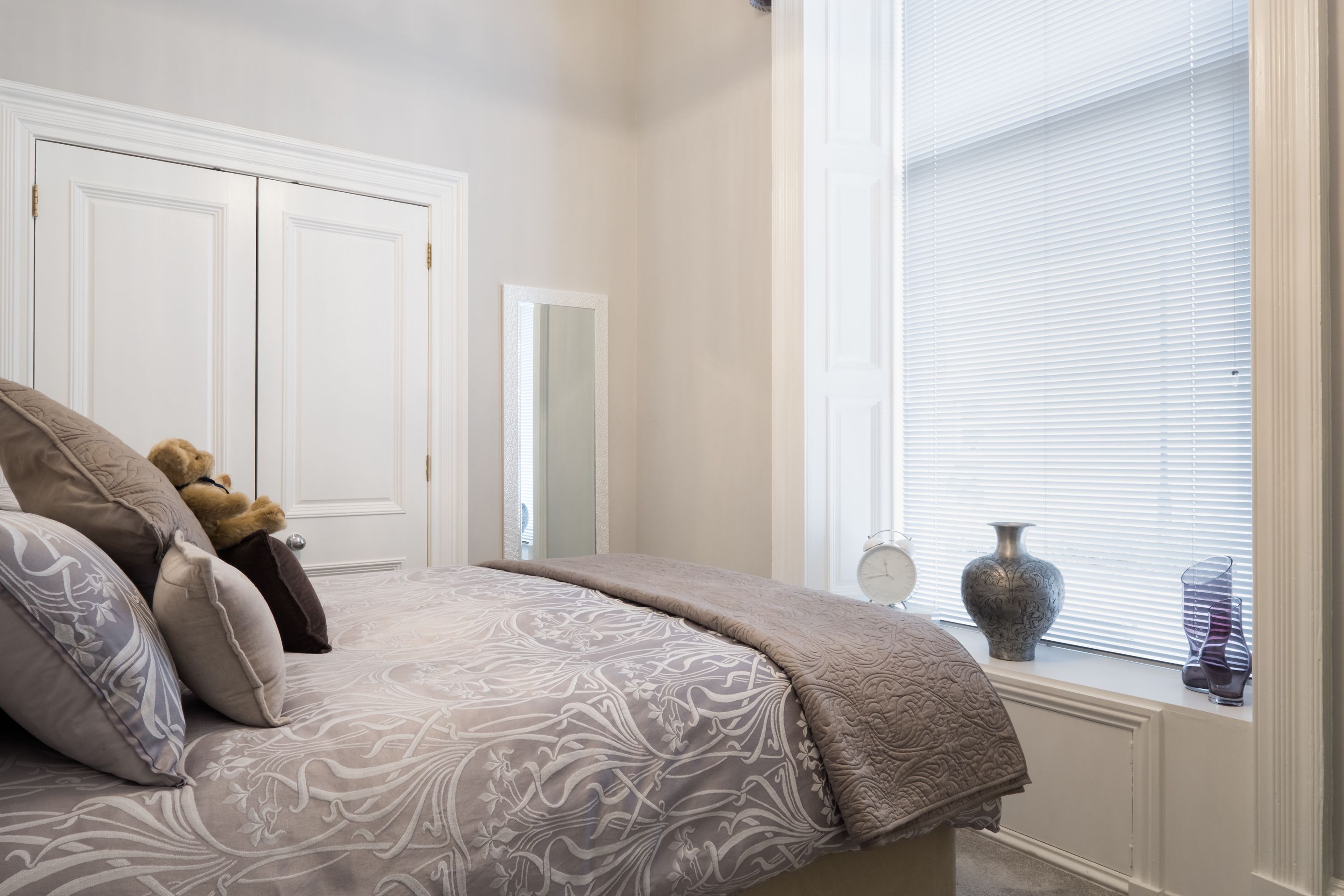 A bedroom with a bed, pillows, a teddy bear, a mirror, and a window with blinds.
