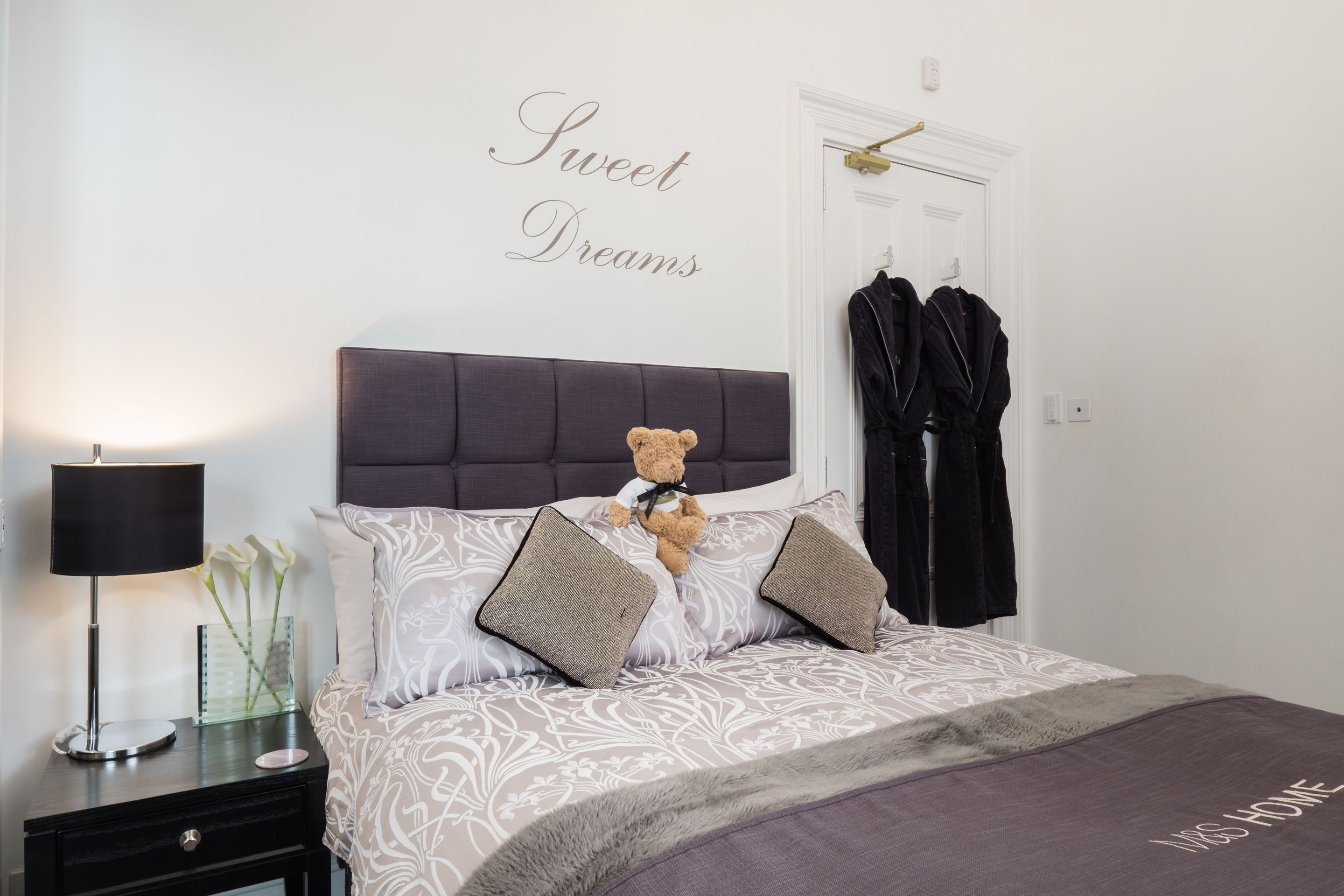 Een net opgemaakt bed met een teddybeer, een bedlampje en een bordje met de tekst 