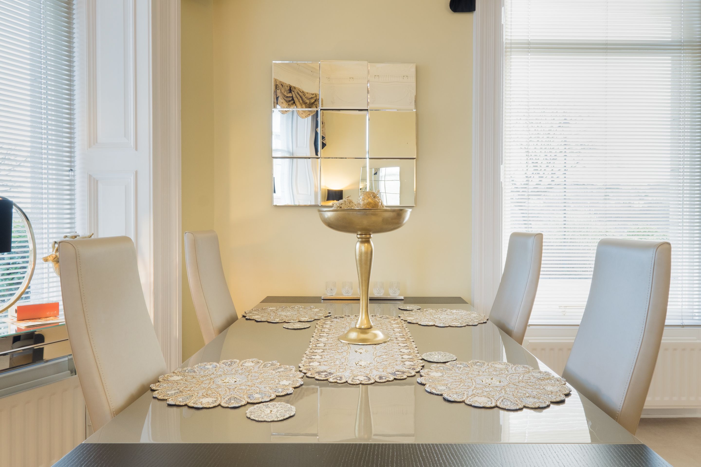 A dining table with a central gold bowl, surrounded by four chairs, and a decorative mirror on the wall.