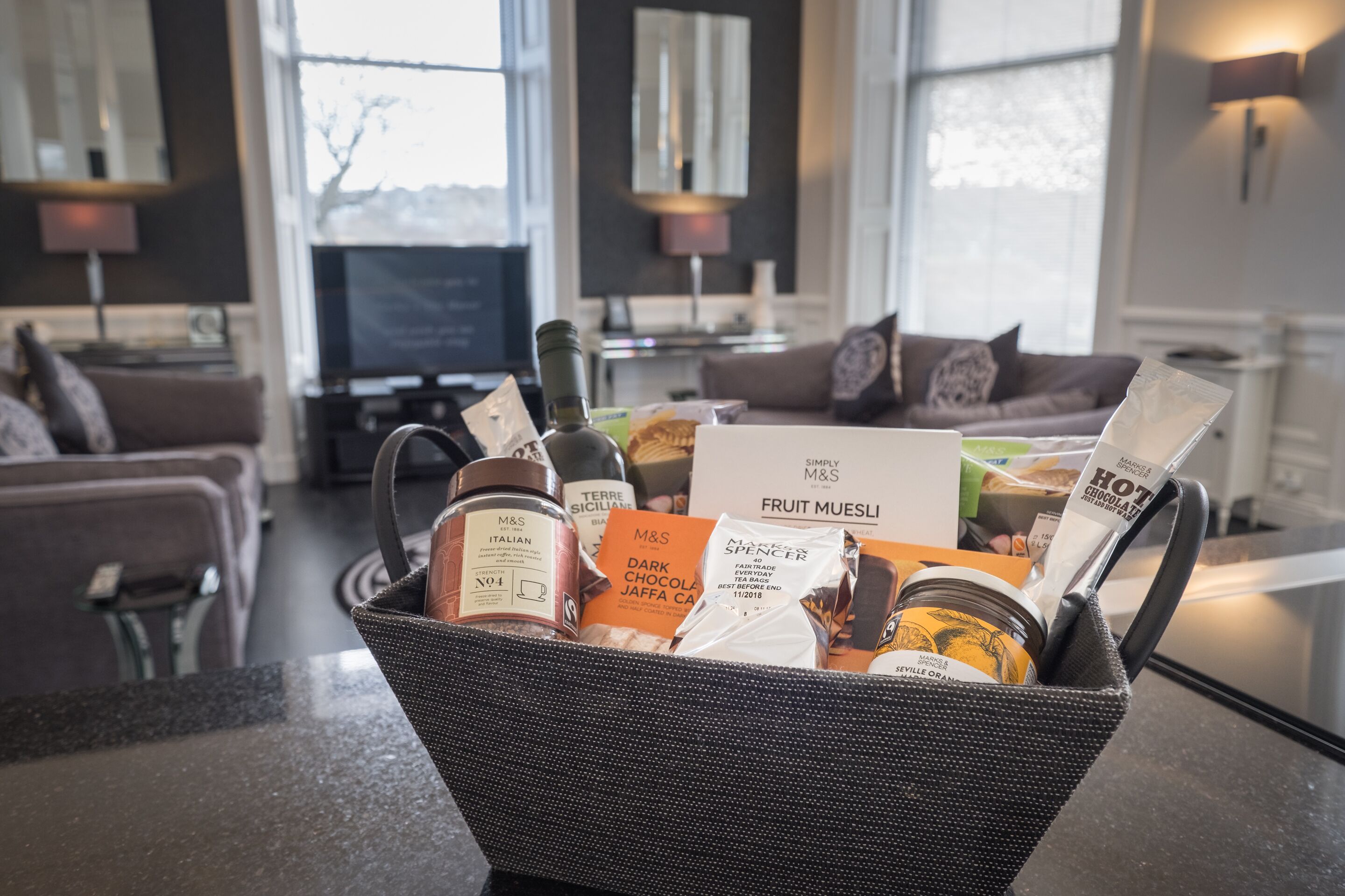 A basket filled with various food items and a bottle of Italian wine in a modern living room with a sofa, TV, and large windows.