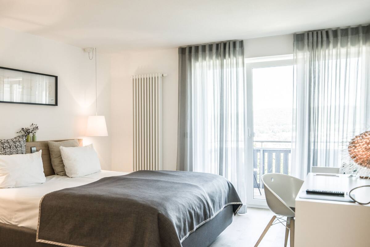A modern hotel room with a large bed, a desk, and a view of the city through the window.