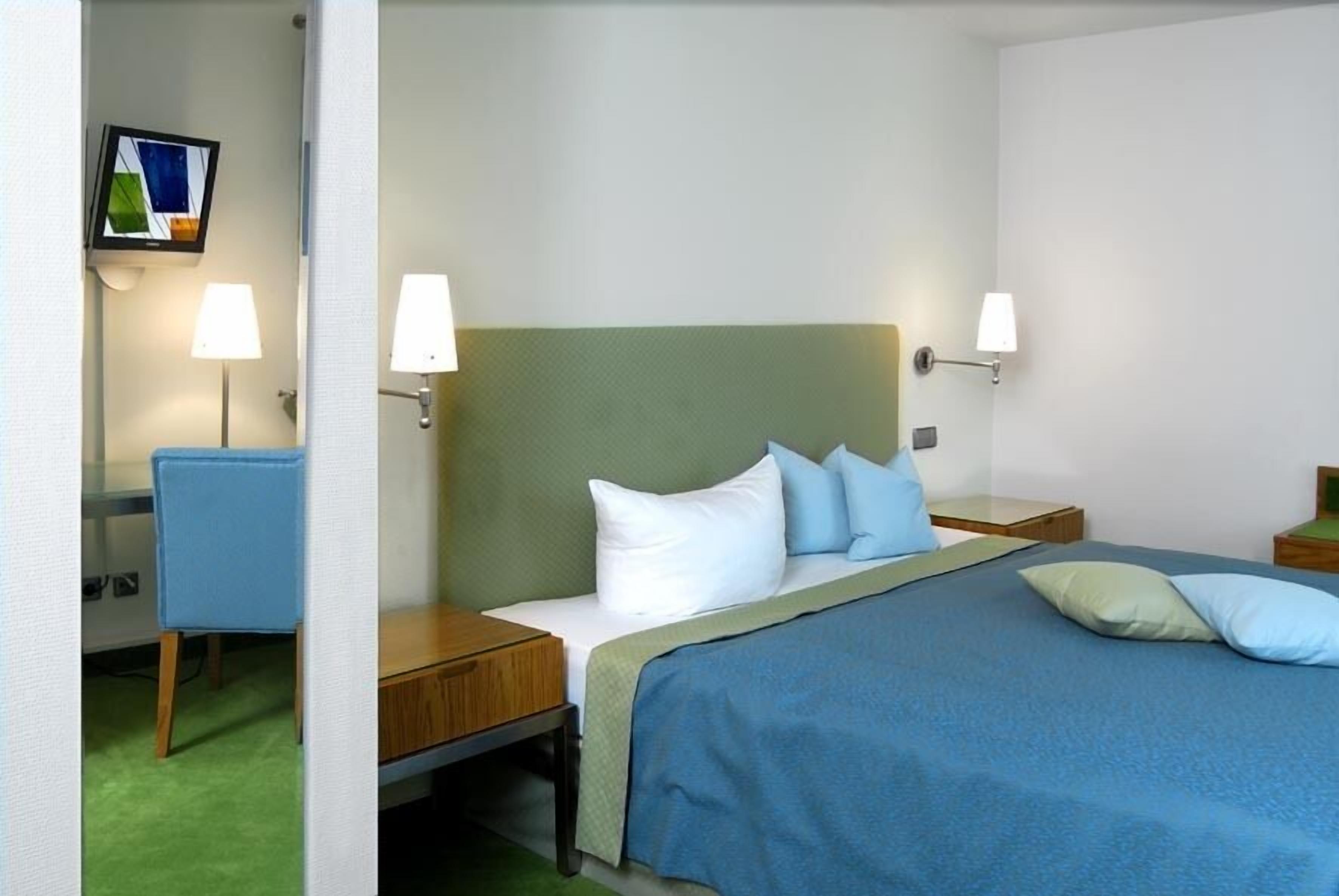 Una habitación de hotel con cama, dos mesitas de noche, una silla, un escritorio y televisor montado en la pared.