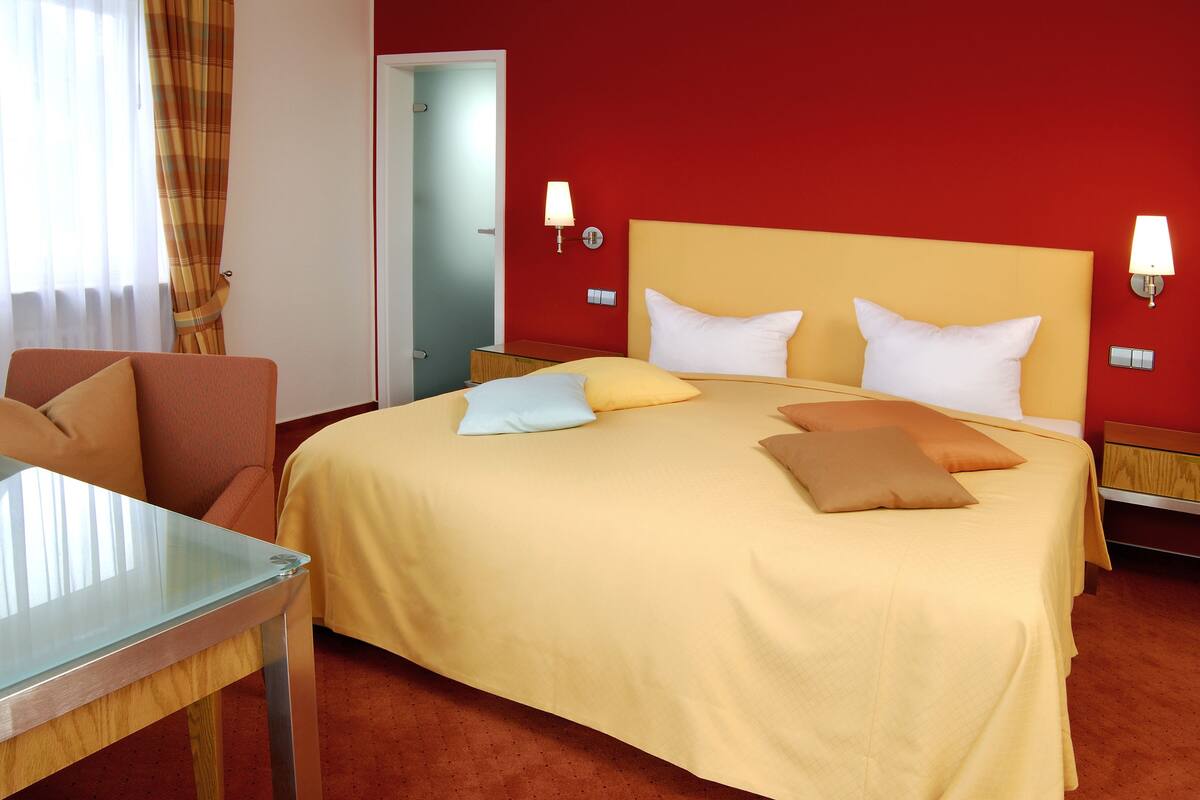 A hotel room with a bed, a chair, a table, and a red wall.