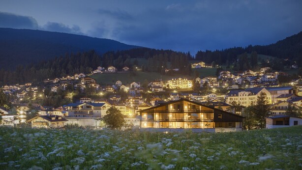 A town at dusk with illuminated buildings, a prominent wooden house in the foreground, and a mountainous backdrop. - Garni Hotel Pra Palmer (Castelrotto)