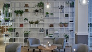 Een modern interieur met een witte wandplank vol planten en boeken, twee grijze fauteuils rond een houten salontafel, en een hanglamp.