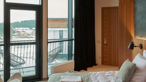 A modern hotel room with a large bed, a sitting area with a chair, a view through large windows, and a ceiling with wooden beams. - Vytautas Mineral SPA (Birstonas)