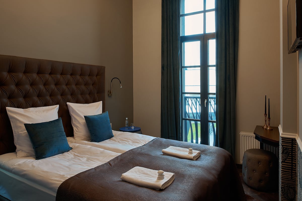 Een hotelkamer met een groot bed, twee kussens, een nachtkastje, een raam met gordijnen en een radiator.