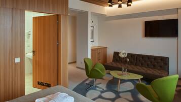 A modern hotel room with a wooden wardrobe, a sofa, two green chairs, a small table with a vase, and a flat-screen TV mounted on the wall.