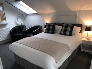 A neatly made bed with a white comforter and two plaid pillows, a black leather chair, a bedside table with a lamp, and a skylight above the bed. - Tirah Bed and Breakfast (Leiston)