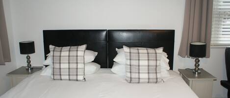 A neatly made bed with a black headboard, two bedside tables with lamps, and a window with blinds.