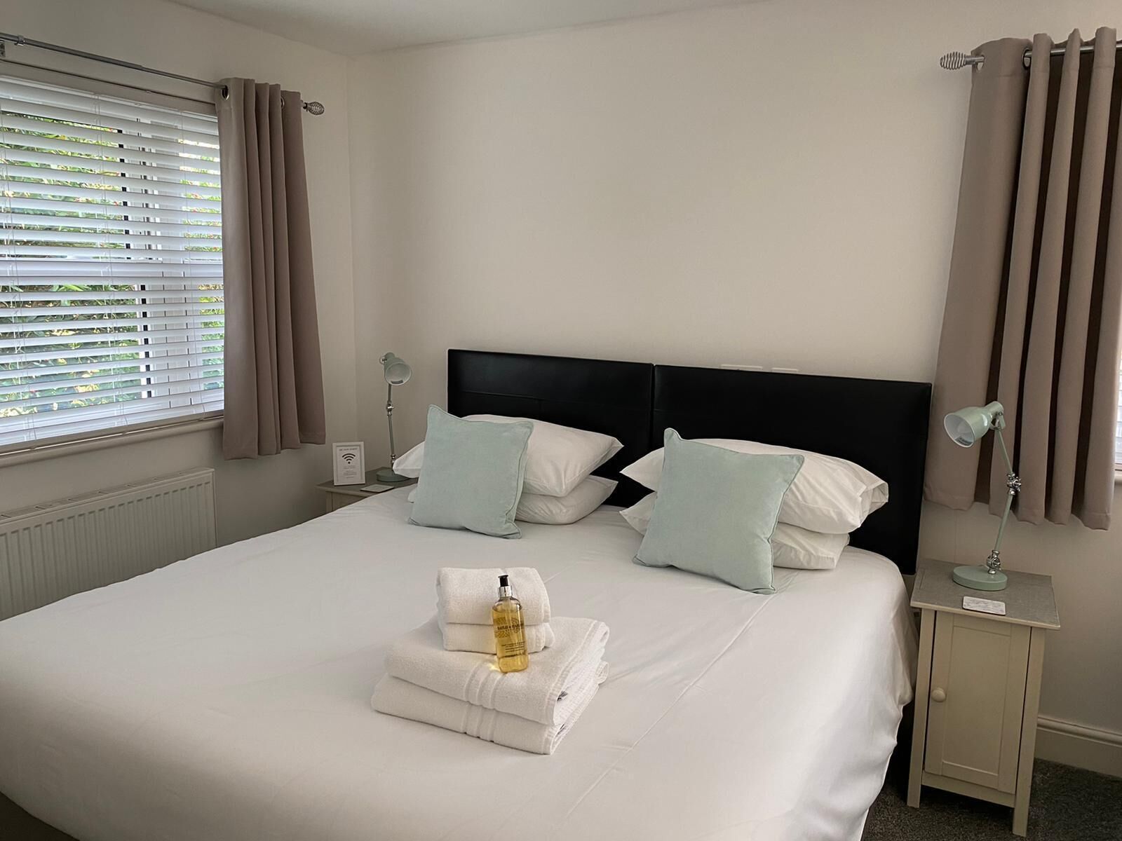 A neatly made bed with white linens, two pillows, and a folded towel with a bottle on it. There is a bedside table with a lamp and a small card.