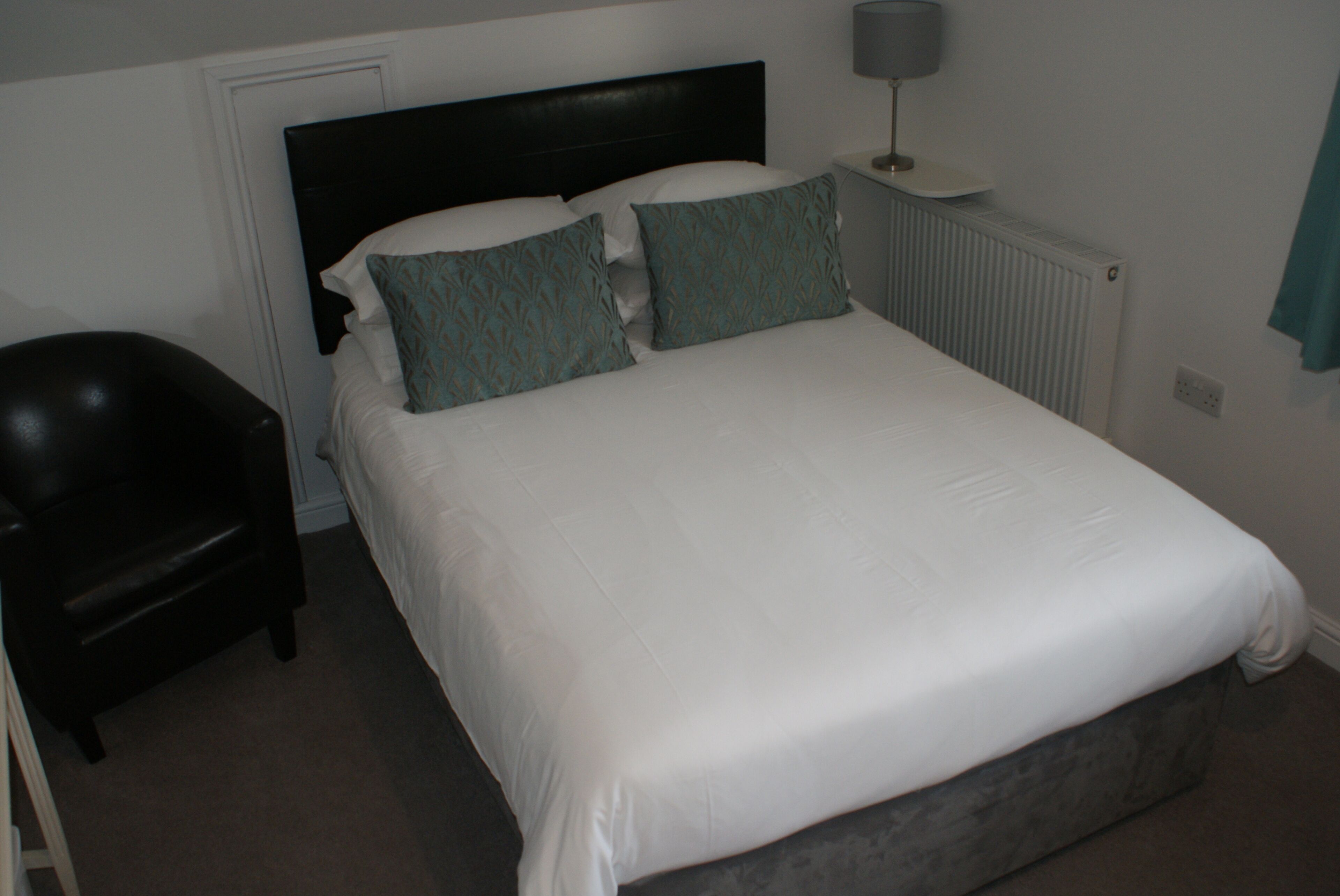 A neatly made bed with white linens and two teal pillows, a black chair, and a bedside table with a lamp.