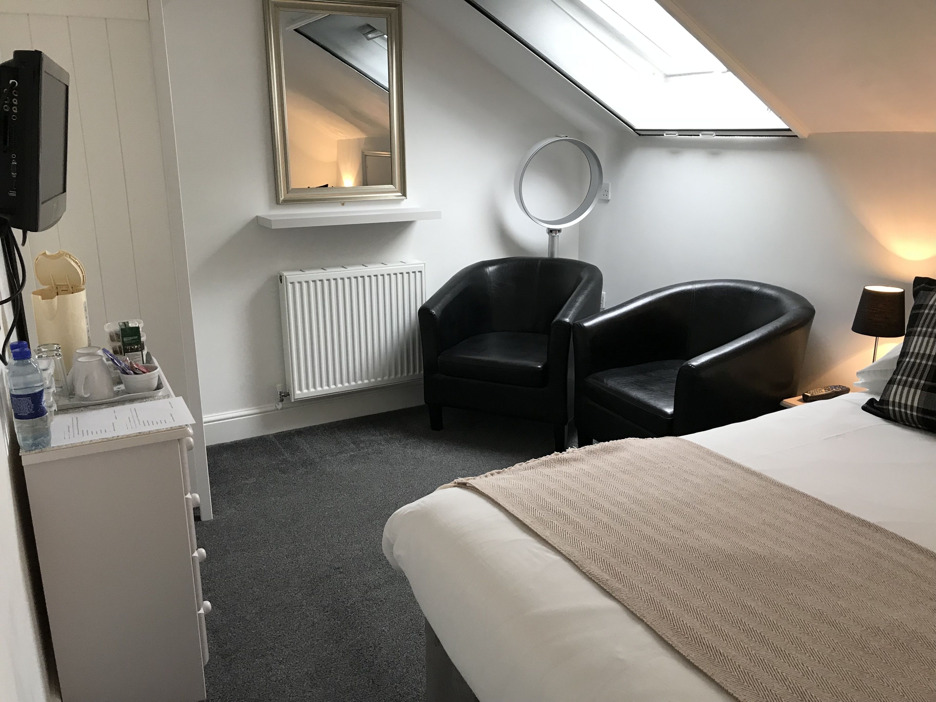 A hotel room with a bed, two black leather chairs, a round mirror, a white radiator, a small table with toiletries, and a skylight.