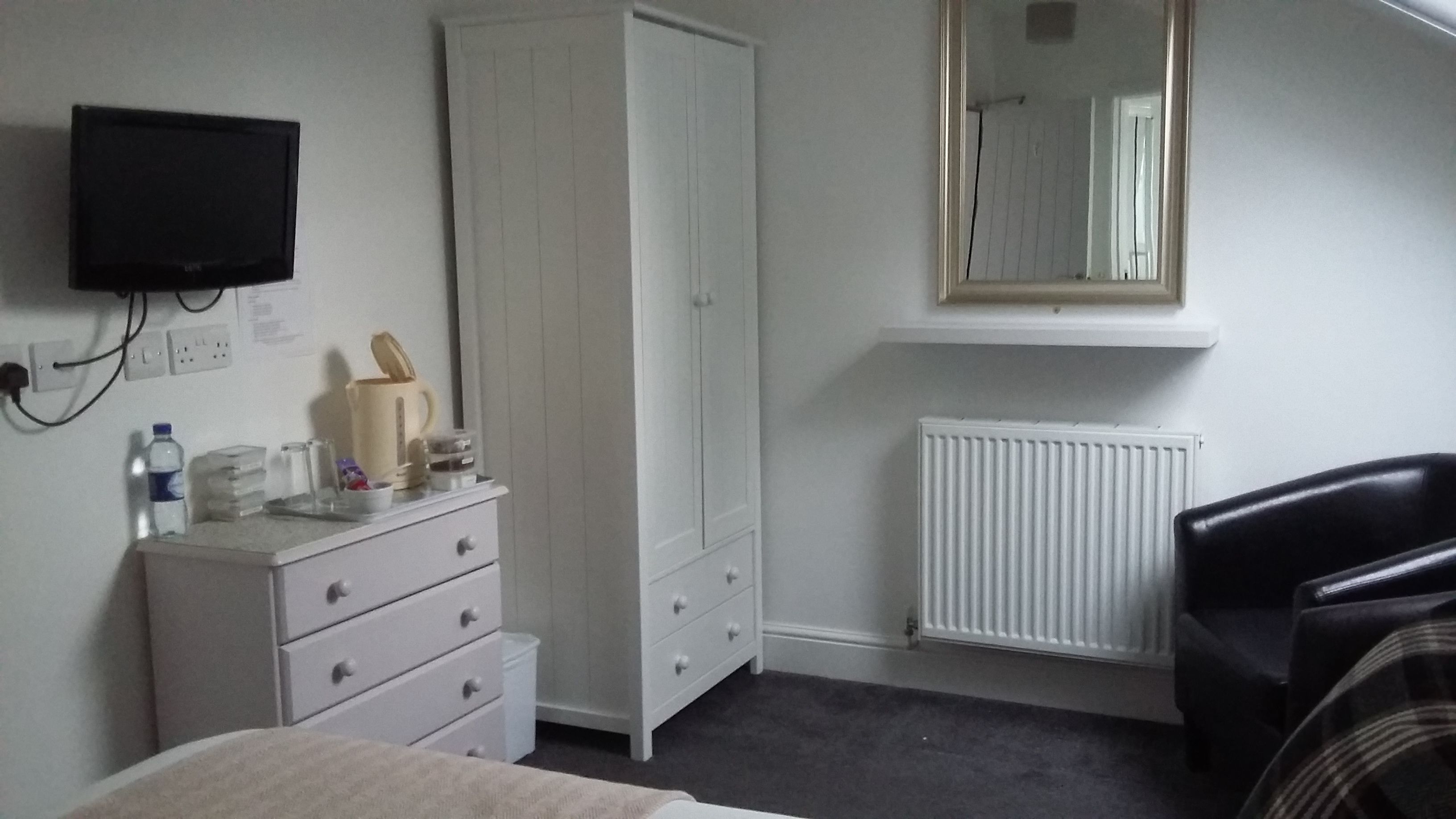 A bedroom with a bed, a white wardrobe, a radiator, a television mounted on the wall, and a chair.
