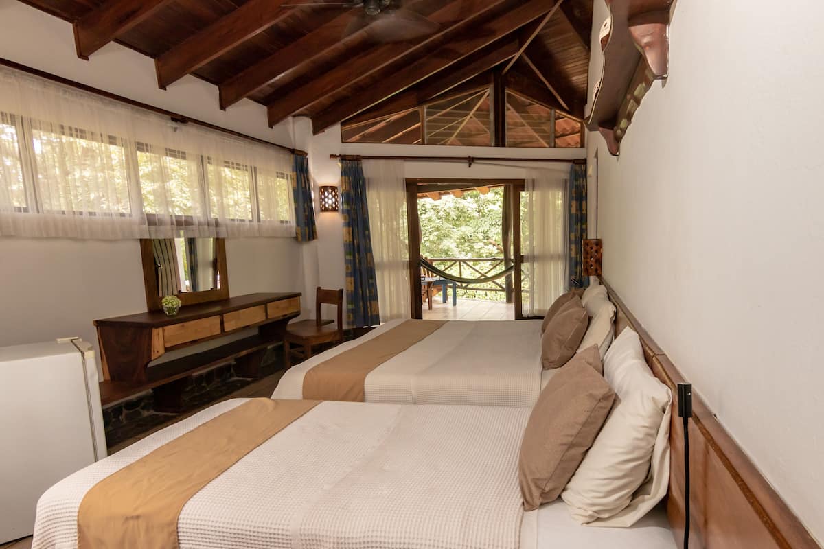 A room with two beds, a wooden bench, a small table, and a view of the outdoors.