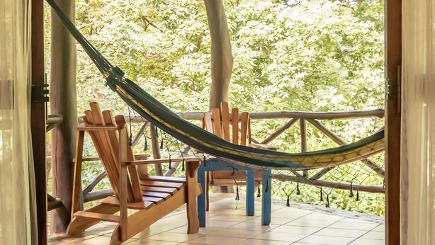Een hangmat onder een houten dak, houten stoelen en een betegelde vloer.