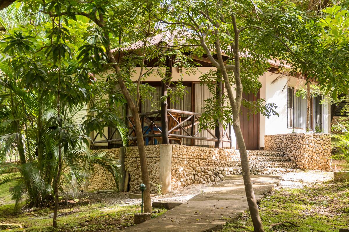 A stone-walled building with a wooden staircase leading to a door, surrounded by lush greenery and a stone pathway.