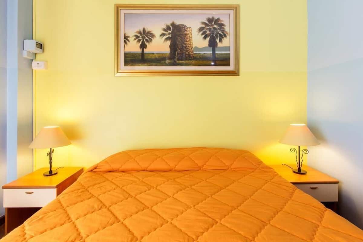 Ein Hotelzimmer mit einem Bett, zwei Nachttischen, einer Lampe auf jedem Tisch, einem Bild an der Wand und einem Thermostat.