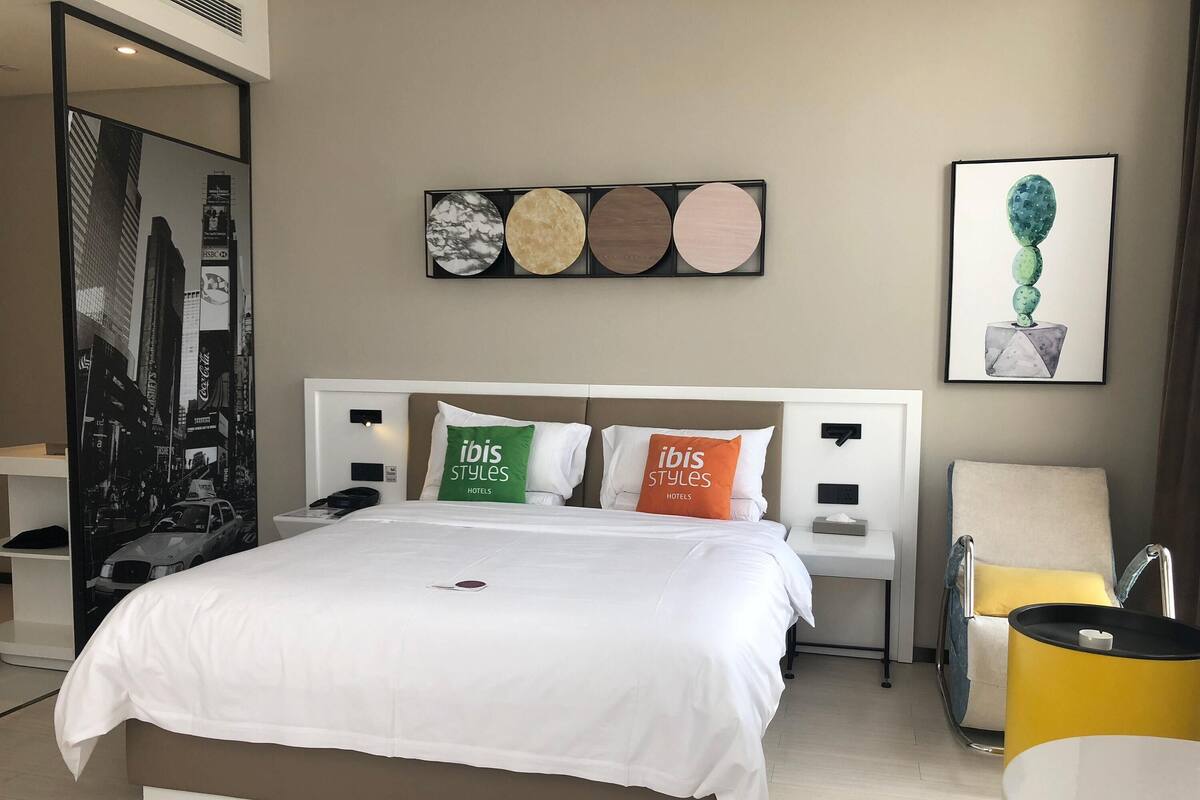 A hotel room with a bed, two nightstands, a chair, and a wall with artwork.