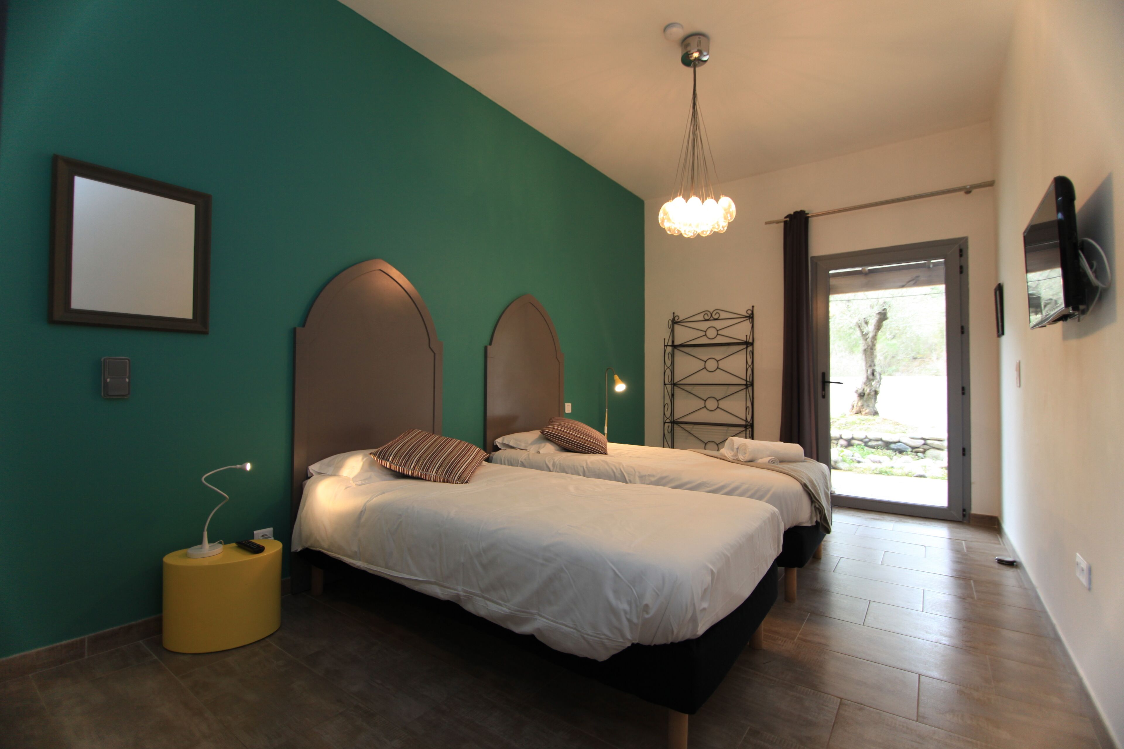 A bedroom with two beds, a yellow side table, a ceiling light, and a TV mounted on the wall.