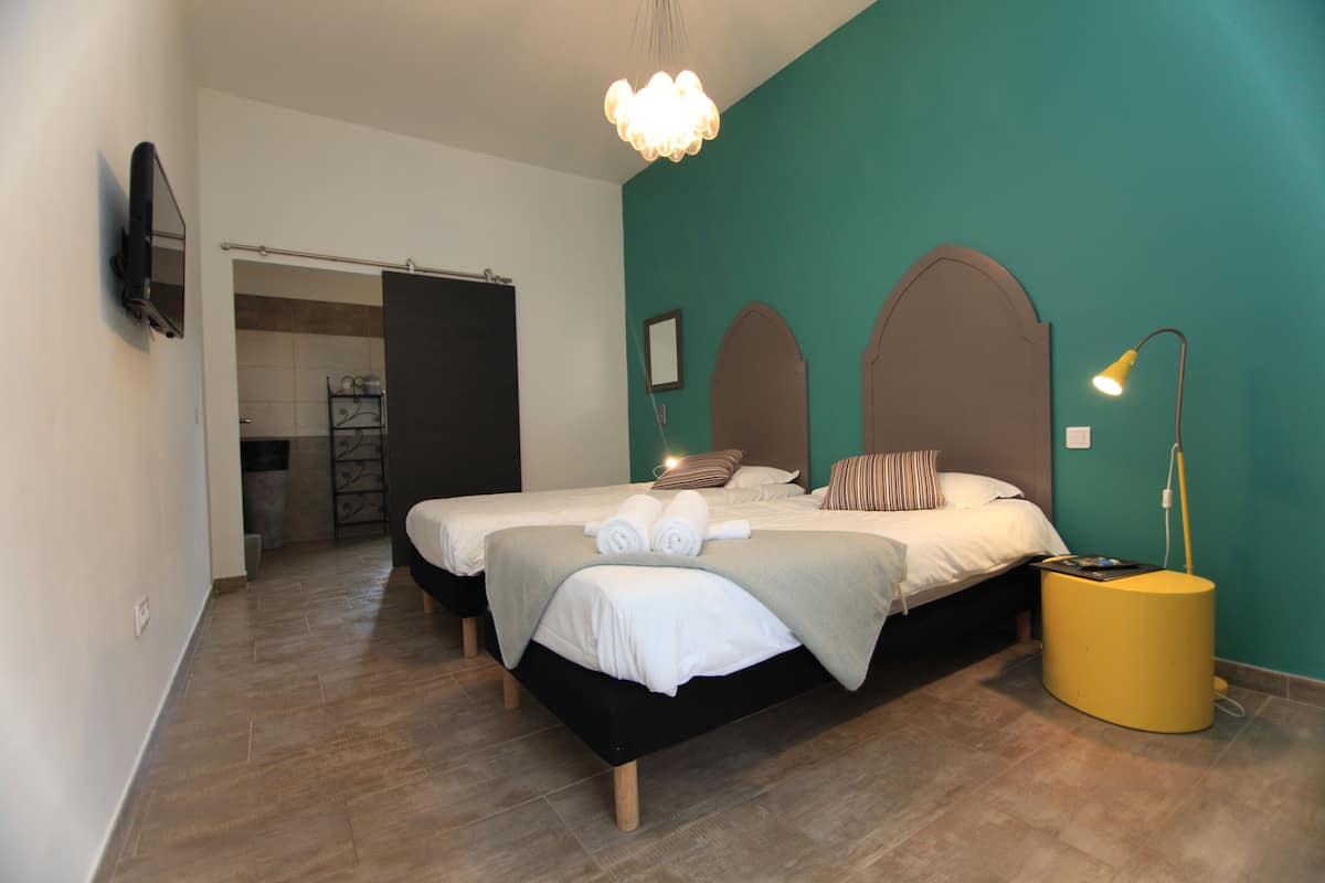 A bedroom with two beds, a TV mounted on the wall, a bedside lamp, and a bathroom visible through an open door.
