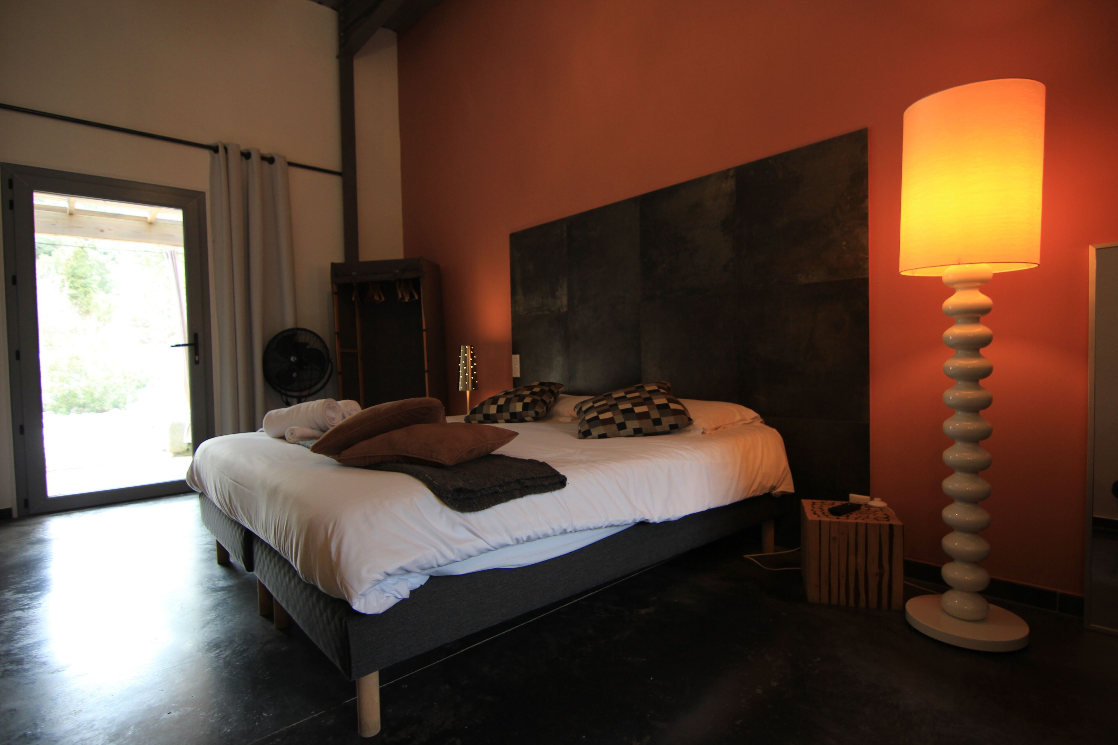 A bedroom with a bed, a standing lamp, a small table, and a door leading to a balcony.