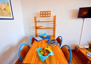 Uma mesa de jantar de madeira com uma cesta de frutas, cadeiras azuis, uma estante de madeira, um quadro emoldurado, uma televisão e um telefone.