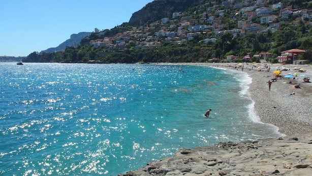 Une plage côtière aux eaux turquoise et limpides, avec un rivage rocheux et des bâtiments construits sur les flancs de la colline.