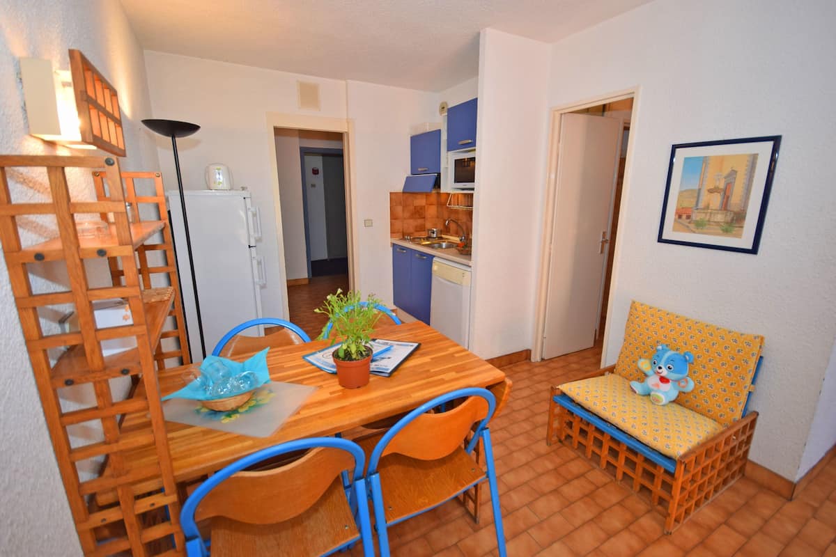 A compact kitchen with a dining area, a small table with chairs, a sofa with a cushion, and a potted plant on the table.