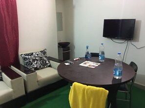 A room with a round table, a sofa with a floral pillow, a small cabinet, a television on the wall, and a green carpet. - AMtec Fazilla Castle (Dhaka)