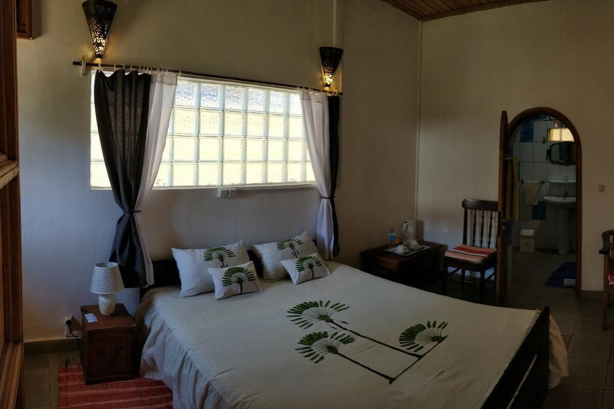 A bedroom with a large bed, a nightstand, a chair, a desk, a window with curtains, a mirror, and a bathroom visible through an archway.