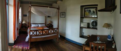 A four-poster bed with a canopy, a desk with a lamp, a shelf with a fan, and a small table with a water bottle and a kettle.