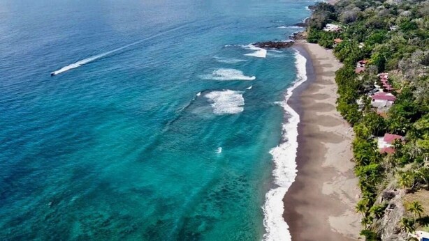 Vista aérea de una playa con aguas turquesas cristalinas, una orilla arenosa y una hilera de casas con tejados rojos.