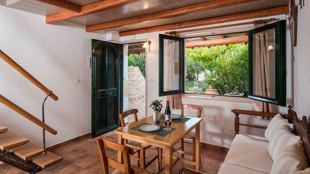 A dining area with a wooden table and chairs, a sofa, and a window with a view of greenery. - Orionas Village (Chania)