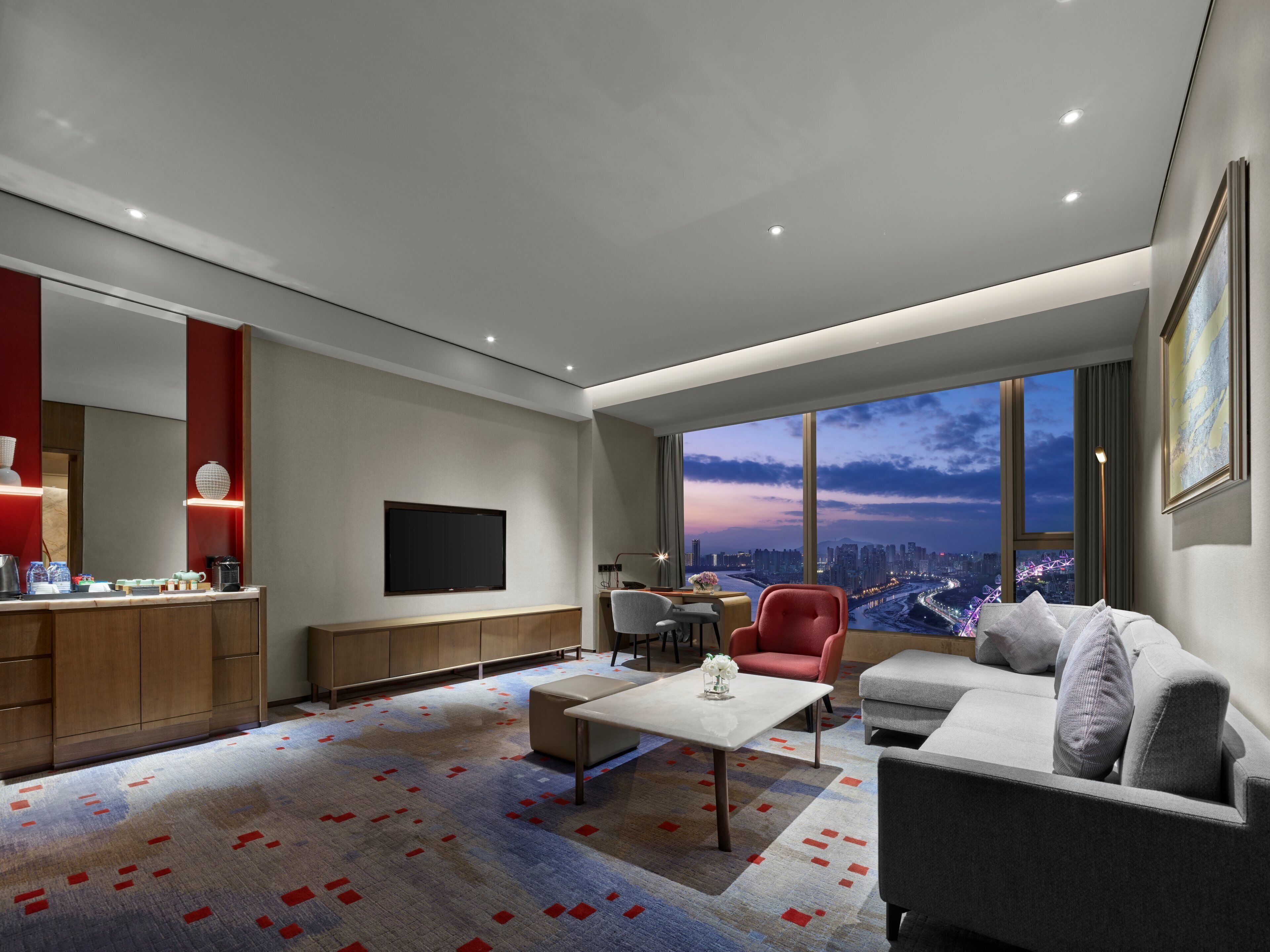 A modern living room with a large window, a flat-screen TV, a coffee station, and a seating area with a sofa and chairs.