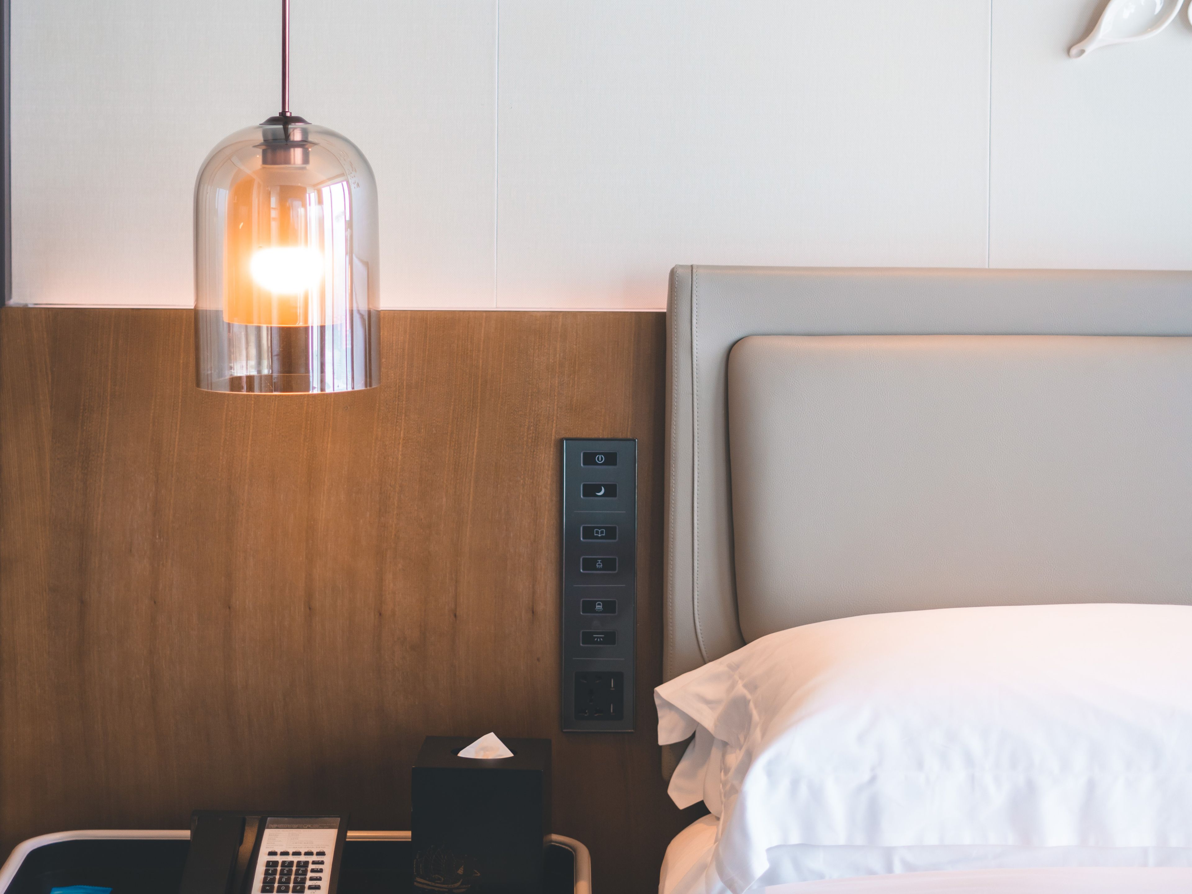 A hotel room with a wooden headboard, a bedside table with a lamp, a telephone, and a power outlet.