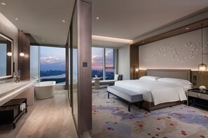 Una habitación de hotel moderna con una cama grande, una zona de estar con vistas, una bañera y un ventanal amplio con vista a la ciudad.