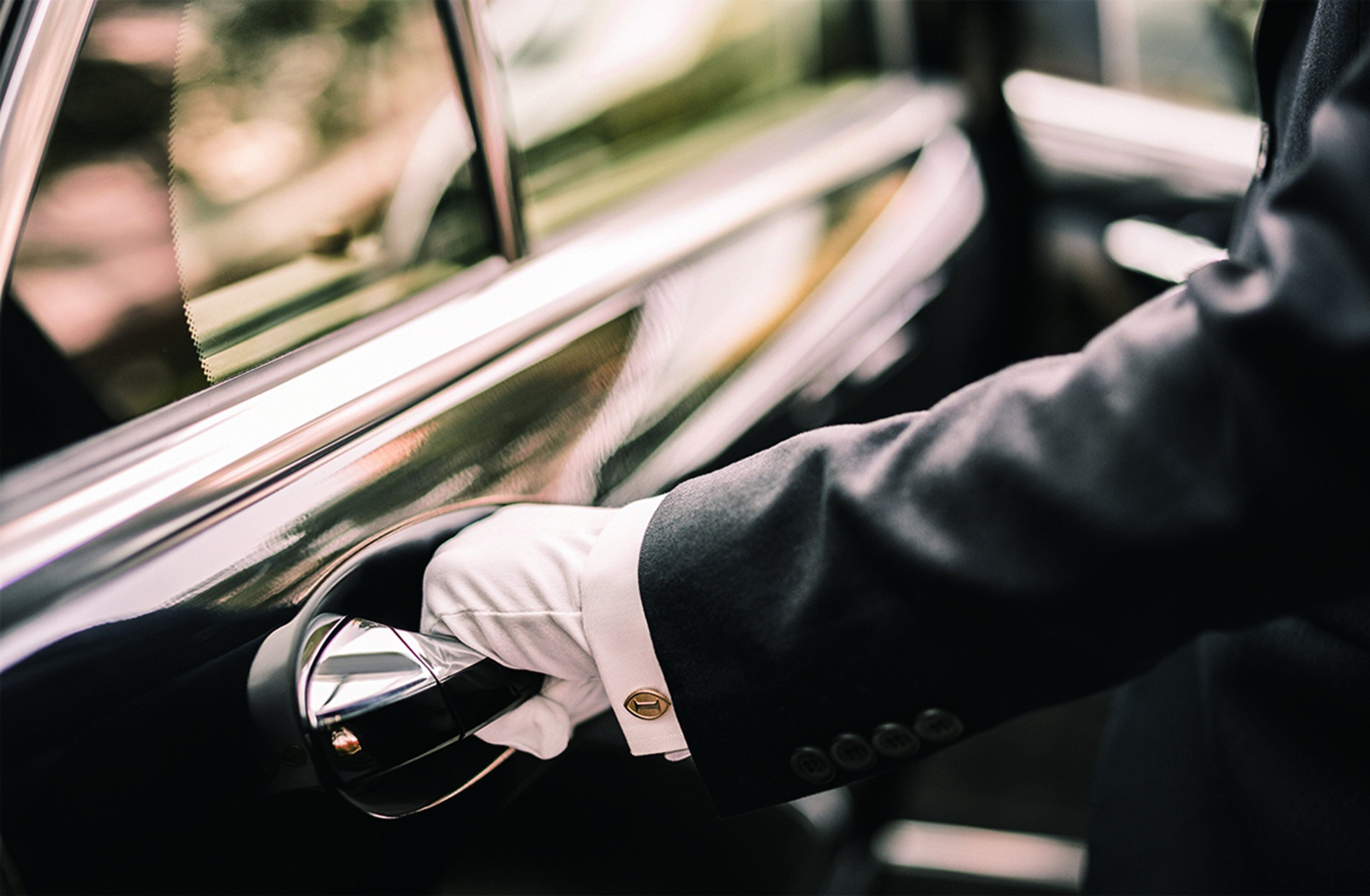 Una persona con guante blanco y traje oscuro, sosteniendo el pomo de una puerta de coche.