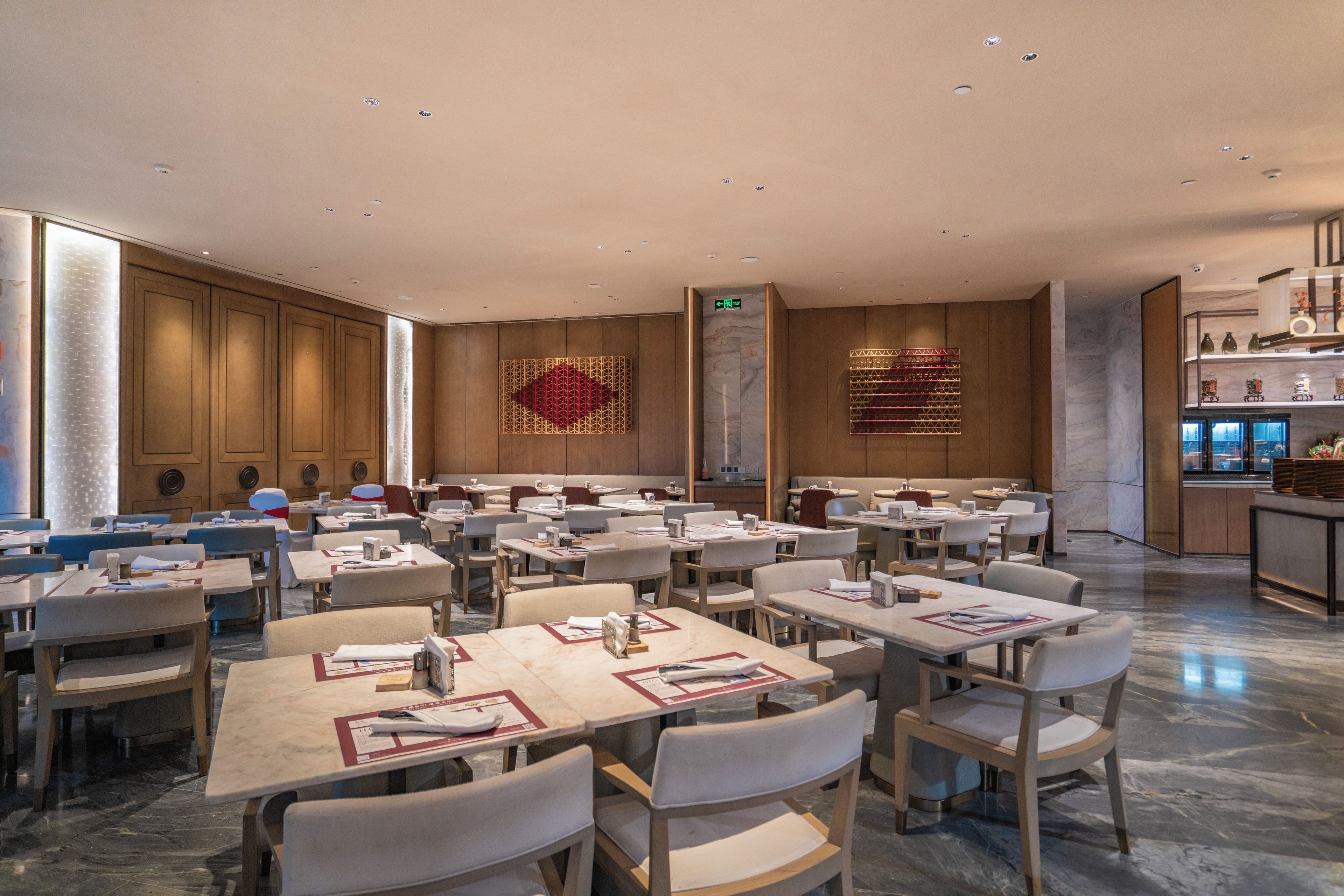 Un vaste espace de restauration d’hôtel, avec de nombreuses tables et chaises, un grand élément décoratif mural et une cuisine moderne visible au fond.