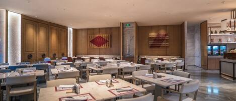 A spacious hotel dining area with multiple tables and chairs, a large decorative wall piece, and a modern kitchen visible in the background.