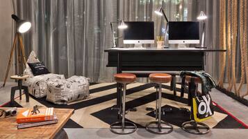 A modern living room with two Apple iMacs on a black desk, a wooden coffee table with books, a bean bag chair, and a bar stool with a bag.