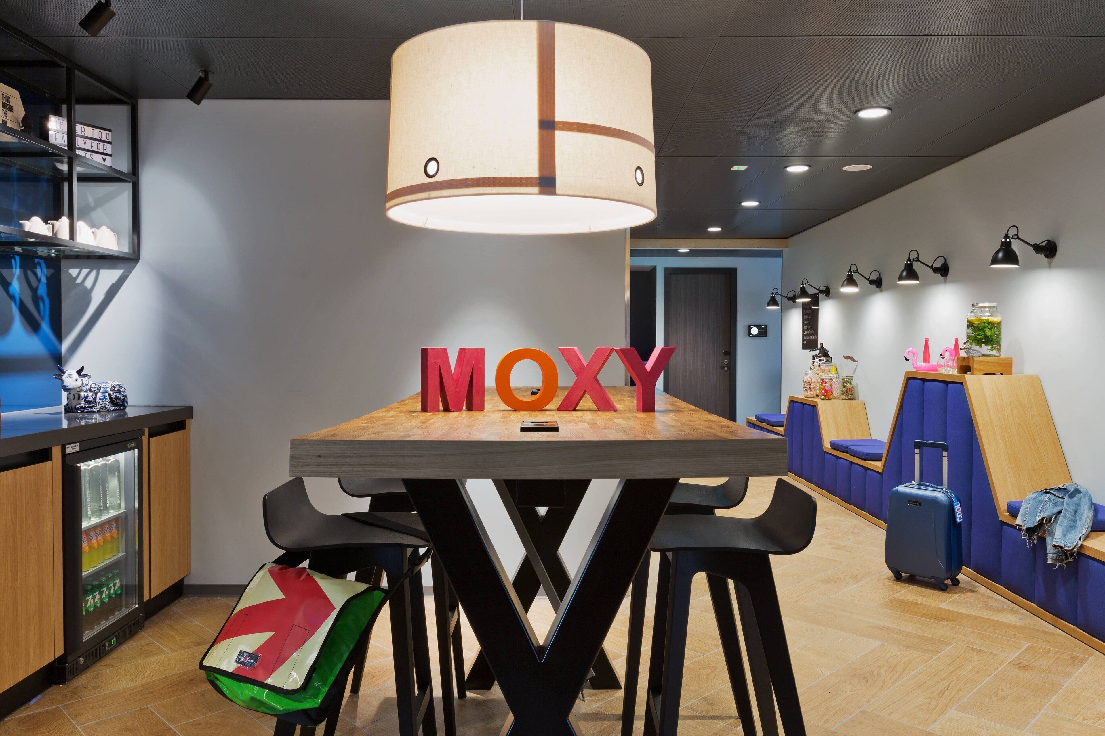 A modern hotel lobby with a central table displaying the word "MOXY", a seating area with a blue sofa, a wooden counter with a refrigerator, and a wall-mounted shelf with decorative items.